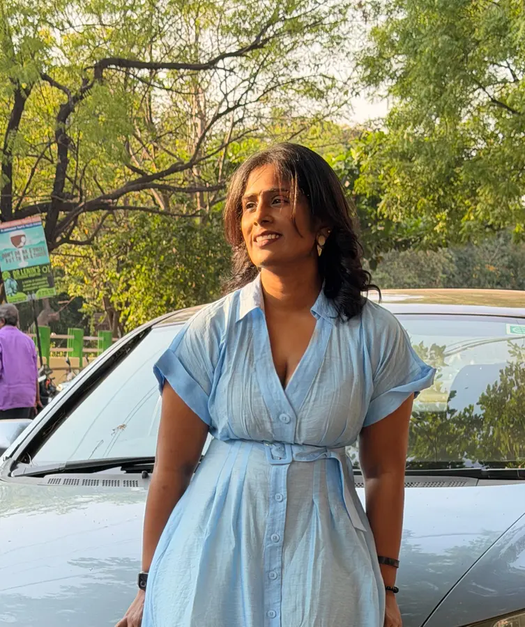 Dr. Indhu Nagaiah in a light blue dress, smiling and looking away, under tall trees