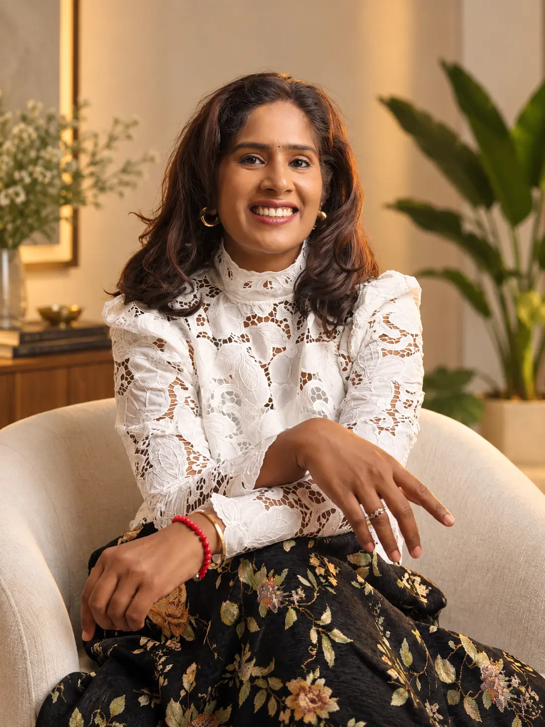Dr. Indhu Nagaiah smiling, seated in a white lace blouse in a softly lit room with plants